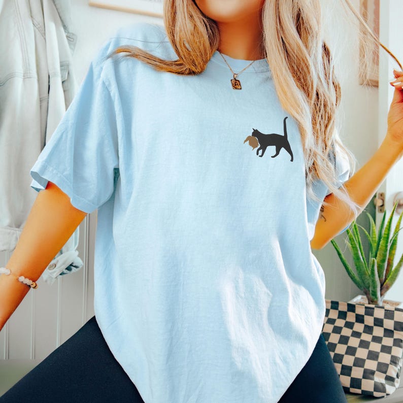 May include: A light blue t-shirt with a black cat carrying a brown object printed on the front.