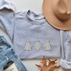 May include: A gray sweatshirt with three embroidered white ghosts on the front. The sweatshirt is laid flat on a white surface with a brown hat and a blue jean jacket.
