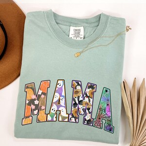 Puede incluir: Una camiseta verde claro con la palabra "MAMA" impresa en letras grandes y coloridas. Las letras est&aacute;n decoradas con varios patrones de tem&aacute;tica de Halloween, incluyendo calabazas, fantasmas, girasoles y telara&ntilde;as.