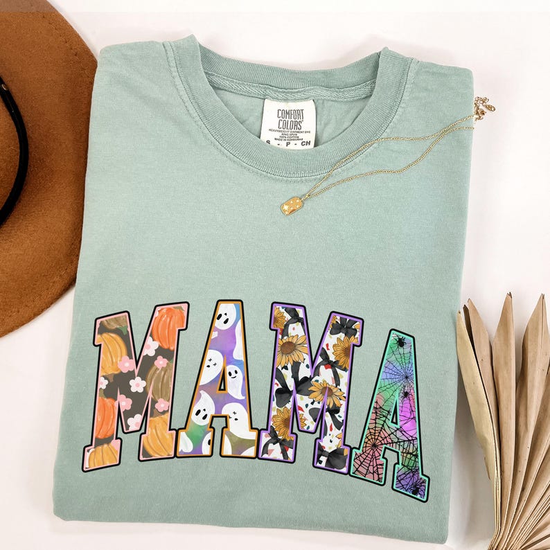 Puede incluir: Una camiseta verde claro con la palabra "MAMA" impresa en letras grandes y coloridas. Las letras est&aacute;n decoradas con varios patrones de tem&aacute;tica de Halloween, incluyendo calabazas, fantasmas, girasoles y telara&ntilde;as.