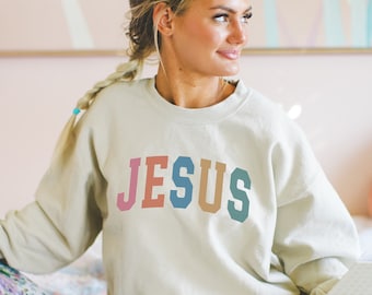May include: A light beige sweatshirt with the word "JESUS" printed in rainbow colors across the front.