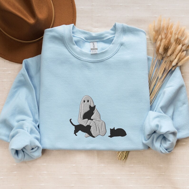 K&ouml;nnte beinhalten: Ein hellblaues Sweatshirt mit einem wei&szlig;en Geist und drei schwarzen Katzen, die auf der Vorderseite gestickt sind. Der Geist sitzt mit einer Katze auf seinem Scho&szlig; und einer weiteren Katze, die neben ihm sitzt. Die dritte Katze liegt vor dem Geist.