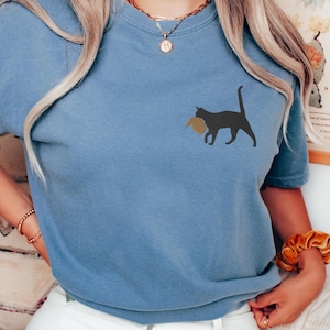 May include: A blue t-shirt with a black cat carrying a brown object in its mouth. The cat is facing the right side of the image.