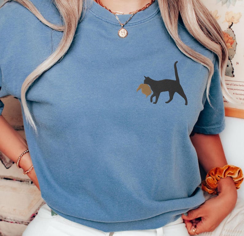 May include: A blue t-shirt with a black cat carrying a brown object in its mouth. The cat is facing the right side of the image.