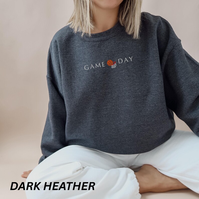 May include: Dark heather gray sweatshirt with the words "GAME DAY" embroidered in white, along with a basketball and hoop graphic. The sweatshirt is a crew neck style and is made of a soft material. The text "DARK HEATHER" is at the bottom of the image.