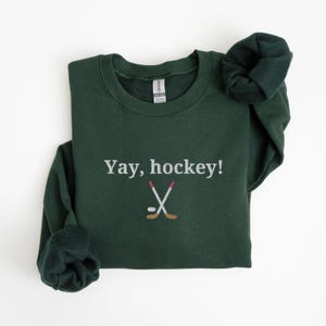 Puede incluir: Una sudadera verde oscuro con el texto "Yay, hockey!" y un gr&aacute;fico blanco de dos palos de hockey cruzados y un disco.