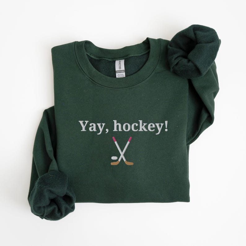 Puede incluir: Una sudadera verde oscuro con el texto "Yay, hockey!" y un gr&aacute;fico blanco de dos palos de hockey cruzados y un disco.