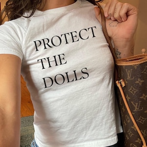 May include: White t-shirt with the text "PROTECT THE DOLLS" in black block letters. The shirt is short-sleeved and appears to be made of a soft material. The person is also carrying a brown handbag with a monogram pattern.
