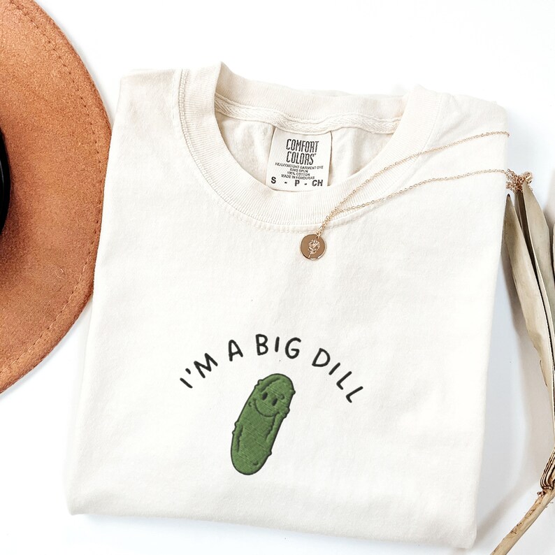 Op de afbeelding: Een wit T-shirt met de tekst "I'm a big dill" en een groene augurk met een smiley. Het shirt is gevouwen en heeft een gouden ketting met een klein hangertje erop.