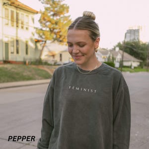 Pu&ograve; includere: Una felpa verde scuro con la scritta "FEMINIST" in lettere bianche. La felpa ha una vestibilit&agrave; ampia e maniche lunghe. La parola "PEPPER" &egrave; stampata in basso a sinistra nell'immagine.