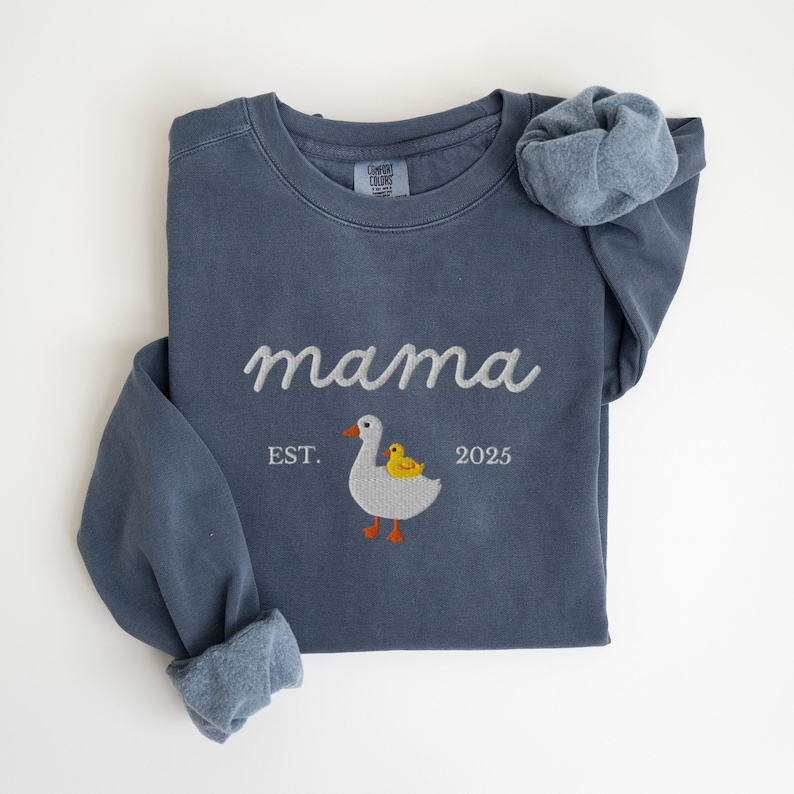 May include: A blue-grey sweatshirt with the word "mama" embroidered in white script. Below the word "mama" is an embroidered design of a goose with a yellow duckling. The text "EST. 2025" is also embroidered on the sweatshirt.