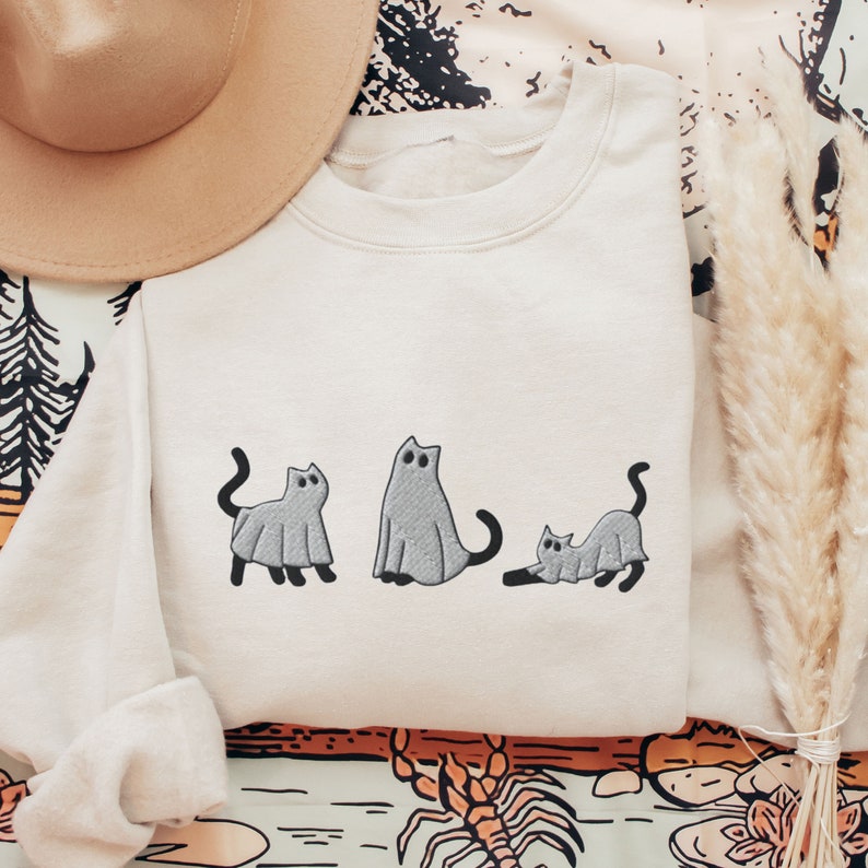 K&ouml;nnte beinhalten: Ein cremefarbener Pullover mit einem Aufdruck von drei grauen Katzen, die als Geister verkleidet sind. Die Katzen stehen in einer Reihe, wobei die mittlere Katze nach vorne und die beiden anderen seitw&auml;rts schauen.