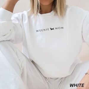 Puede incluir: Sudadera blanca con el texto "WEENIE MOM" en negro y una silueta de perro salchicha. La imagen tambi&eacute;n muestra la palabra "WHITE" en la parte inferior, indicando el color del conjunto.
