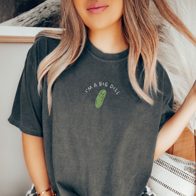 Op de afbeelding: Een donkergrijs T-shirt met de tekst "I'm a big dill" en een groene augurk met een glimlach.