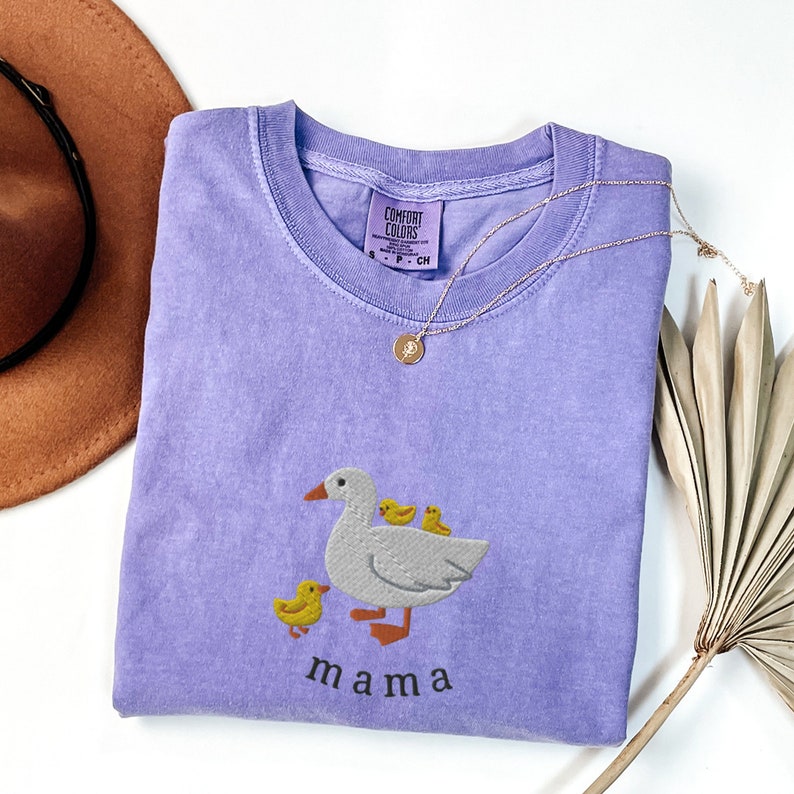 May include: A lavender t-shirt with a white embroidered goose and three yellow ducklings. The text "mama" is embroidered below the goose.