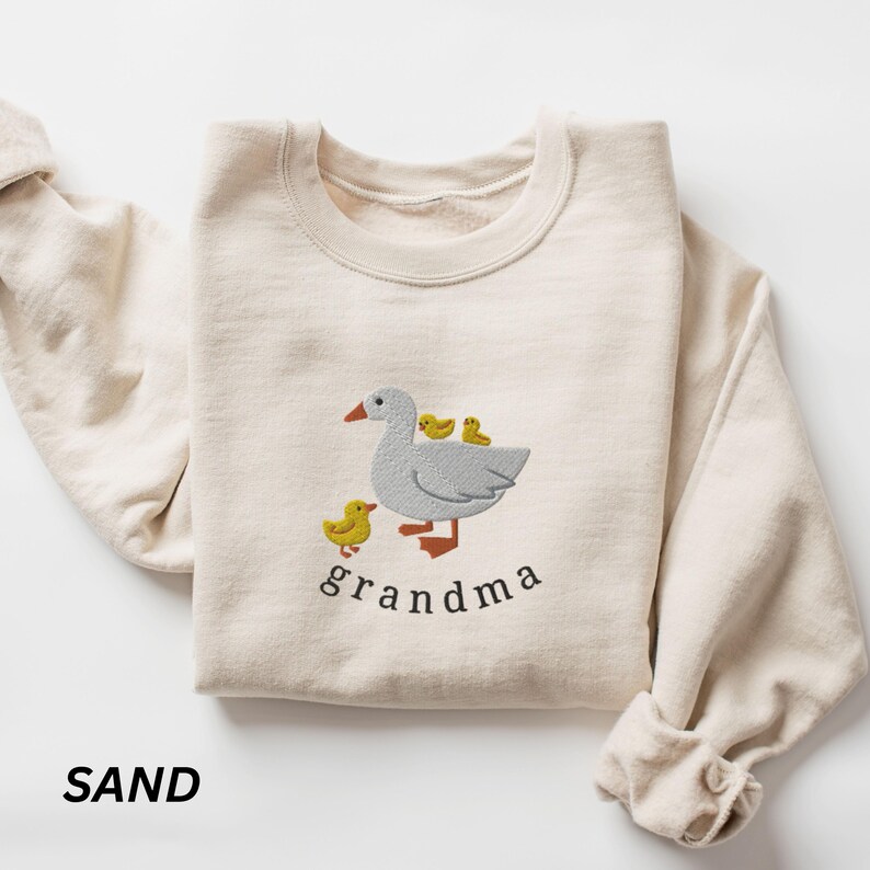 May include: A sand-colored sweatshirt with a white goose and three yellow ducklings embroidered on the front. The text "grandma" is embroidered below the goose.