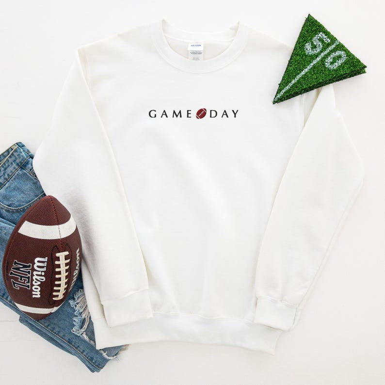 Może przedstawiać: Biała bluza z napisem "GAME DAY" w kolorze czarnym i grafiką piłki nożnej. Brązowa i biała piłka nożna oraz zielony słupek bramki r&oacute;wnież znajdują się na zdjęciu. Bluza jest na parze dżins&oacute;w.