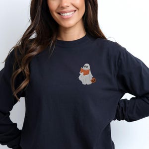K&ouml;nnte beinhalten: Ein schwarzer Langarmsweatshirt mit einem wei&szlig;en Geist, der ein Buch liest und einen kleinen orangefarbenen K&uuml;rbis h&auml;lt. Der Geist ist auf der linken Brust des Sweatshirts gestickt.