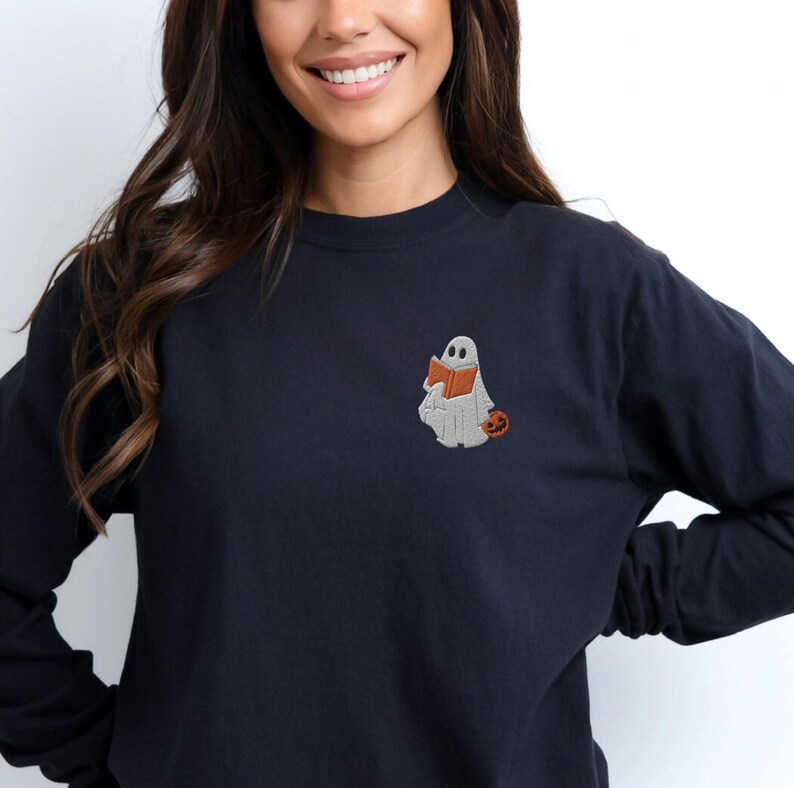 K&ouml;nnte beinhalten: Ein schwarzer Langarmsweatshirt mit einem wei&szlig;en Geist, der ein Buch liest und einen kleinen orangefarbenen K&uuml;rbis h&auml;lt. Der Geist ist auf der linken Brust des Sweatshirts gestickt.