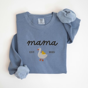 May include: A blue sweatshirt with the word "mama" embroidered in black script. Below the word "mama" is an embroidered image of a goose and a duckling. The text "EST. 2025" is below the goose and duckling.