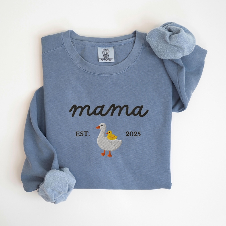 May include: A blue sweatshirt with the word "mama" embroidered in black script. Below the word "mama" is an embroidered image of a goose and a duckling. The text "EST. 2025" is below the goose and duckling.