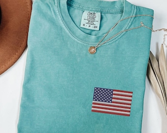 May include: A teal blue t-shirt with a small American flag embroidered on the front. The shirt has a distressed look and is folded in half. The shirt has a tag that says "Comfort Colors" and "S - P - CH".