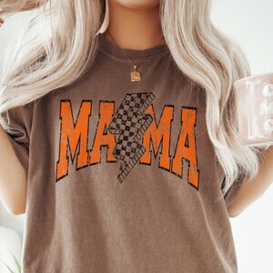 K&ouml;nnte beinhalten: Ein braunes T-Shirt mit einer orangefarbenen Grafik des Wortes "MAMA" mit einem karierten Blitz-Design.