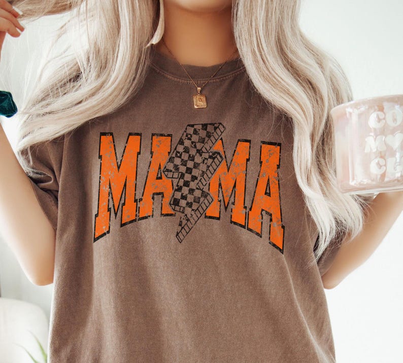 K&ouml;nnte beinhalten: Ein braunes T-Shirt mit einer orangefarbenen Grafik des Wortes "MAMA" mit einem karierten Blitz-Design.