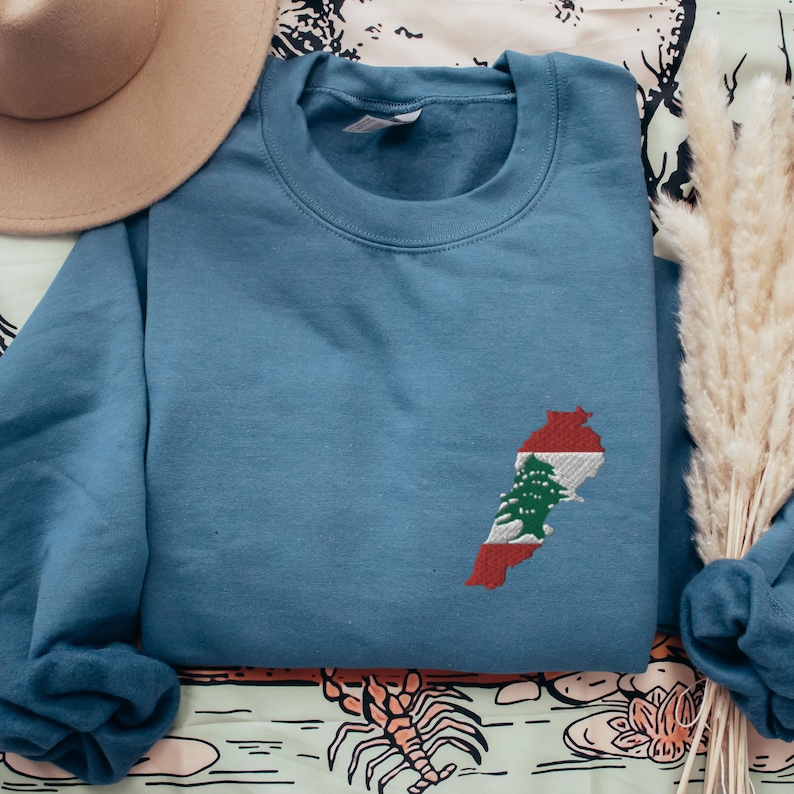 May include: A blue sweatshirt with an embroidered outline of Lebanon with the Lebanese flag colors inside. The flag features a green cedar tree on a white background with red stripes on either side.
