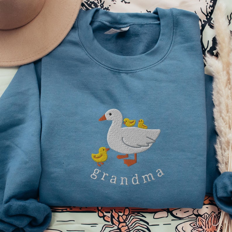May include: A blue sweatshirt with a white embroidered goose and three yellow ducklings. The text "grandma" is embroidered below the goose.