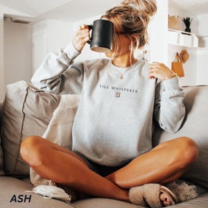 May include: A light gray sweatshirt with the text "TILE WHISPERER" in black. The sweatshirt has a crew neck and long sleeves. A black mug is held up to the face. The word "ASH" is printed in the bottom left corner.