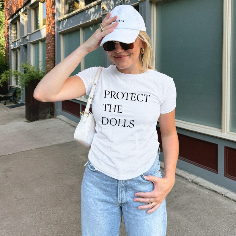 May include: White t-shirt with the text "PROTECT THE DOLLS" in black. The person is wearing a white baseball cap and sunglasses. The person is also wearing light blue jeans and carrying a white purse.