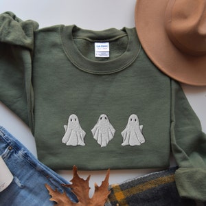 May include: A green sweatshirt with three white embroidered ghosts on the front. The sweatshirt is laid flat on a white surface.