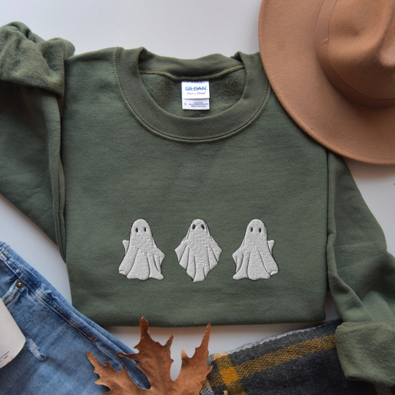 May include: A green sweatshirt with three white embroidered ghosts on the front. The sweatshirt is laid flat on a white surface.