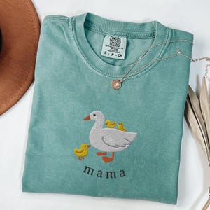 Cute Mama Goose Embroidered with Baby Chicks on Unisex Light Green Comfort Colors T-Shirt, folded with gold necklace