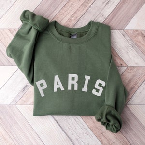 Puede incluir: Una sudadera verde con la palabra "PARIS" impresa en blanco en la parte delantera.