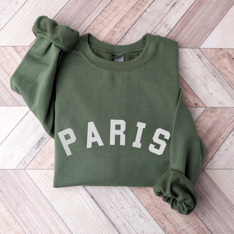 Puede incluir: Una sudadera verde con la palabra "PARIS" impresa en blanco en la parte delantera.