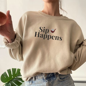 May include: A beige sweatshirt with the text "Sip Happens" and a small graphic of a wine glass with red wine inside.