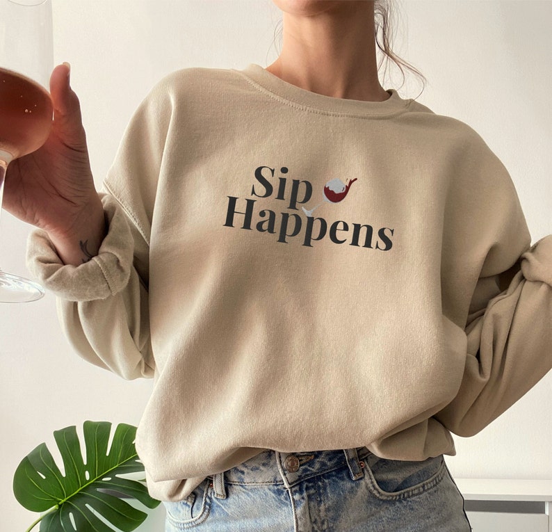 May include: A beige sweatshirt with the text "Sip Happens" and a small graphic of a wine glass with red wine inside.