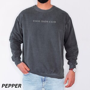 May include: Pepper-colored crewneck sweatshirt with the text "COOL DADS CLUB" in white. The sweatshirt has a vintage, washed look and long sleeves. The model is wearing blue jeans and a watch.