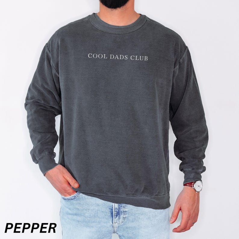 May include: Pepper-colored crewneck sweatshirt with the text "COOL DADS CLUB" in white. The sweatshirt has a vintage, washed look and long sleeves. The model is wearing blue jeans and a watch.