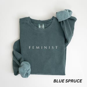 Pu&ograve; includere: Felpa blu abete con la scritta "FEMINIST" stampata in bianco. La felpa ha un girocollo e maniche lunghe con polsini ripiegati. L'etichetta recita "COMFORT COLORS". L'angolo in basso a destra dell'immagine recita "BLUE SPRUCE".