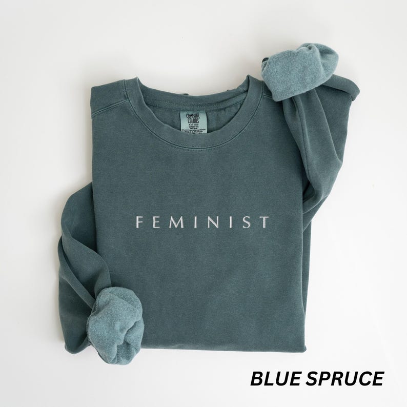Pu&ograve; includere: Felpa blu abete con la scritta "FEMINIST" stampata in bianco. La felpa ha un girocollo e maniche lunghe con polsini ripiegati. L'etichetta recita "COMFORT COLORS". L'angolo in basso a destra dell'immagine recita "BLUE SPRUCE".