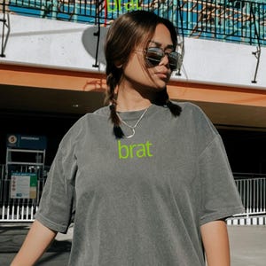 May include: A gray t-shirt with a green "brat" graphic printed on the front. The shirt has a relaxed fit and short sleeves.
