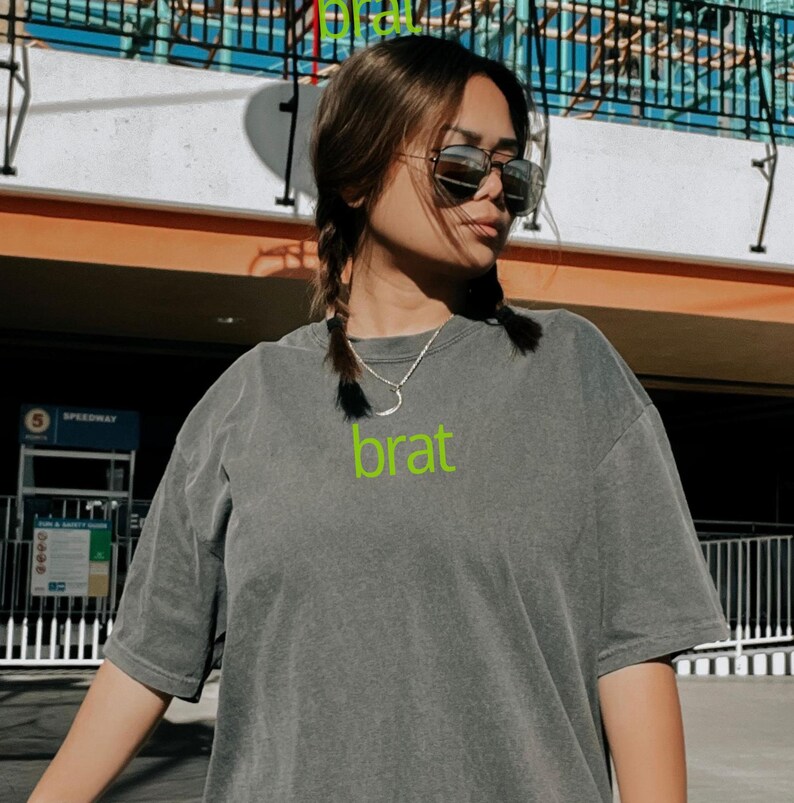 May include: A gray t-shirt with a green "brat" graphic printed on the front. The shirt has a relaxed fit and short sleeves.