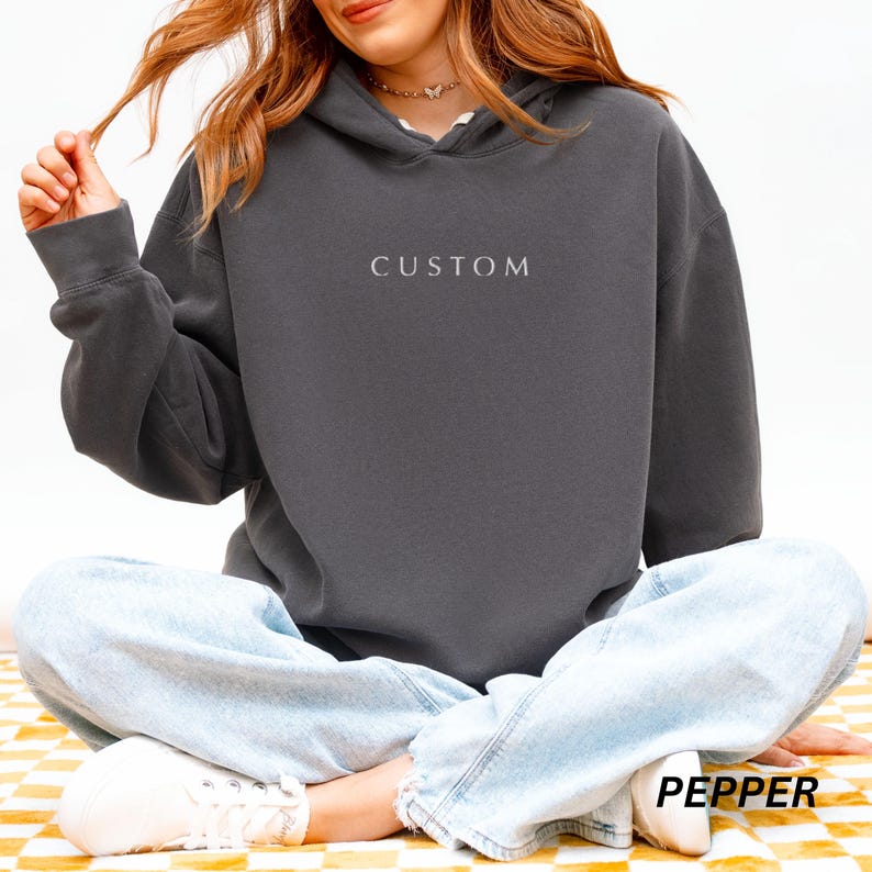 May include: A dark gray hoodie with the word "CUSTOM" embroidered on the front. The hoodie has a drawstring hood and long sleeves. The word "PEPPER" is printed at the bottom right corner of the image.