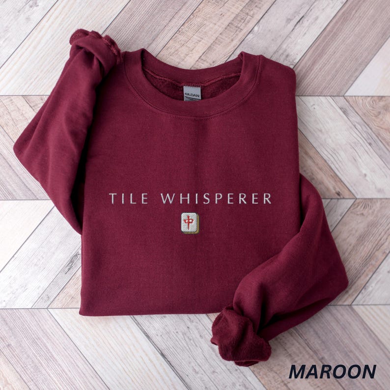 May include: Maroon-colored sweatshirt with the text "TILE WHISPERER" in white. A small, square tile with Chinese characters is below the text. The word "MAROON" is at the bottom right corner of the image.