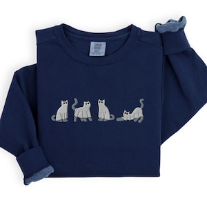 May include: Navy blue sweatshirt with embroidered gray cat design. The cats are in various poses, creating a playful and whimsical look. The sweatshirt has a crew neck and long sleeves with denim-like cuffs.