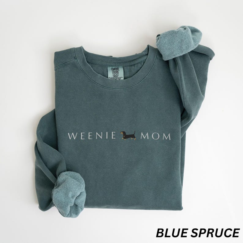 Puede incluir: Sudadera azul abeto con el texto "WEENIE MOM" y una ilustraci&oacute;n de perro salchicha. La sudadera tiene cuello redondo y mangas largas con pu&ntilde;os. El texto "BLUE SPRUCE" est&aacute; en la parte inferior de la imagen.