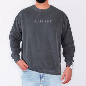 May include: A dark grey sweatshirt with a white embroidered word "HUSBAND" on the front. The sweatshirt has a relaxed fit and long sleeves.